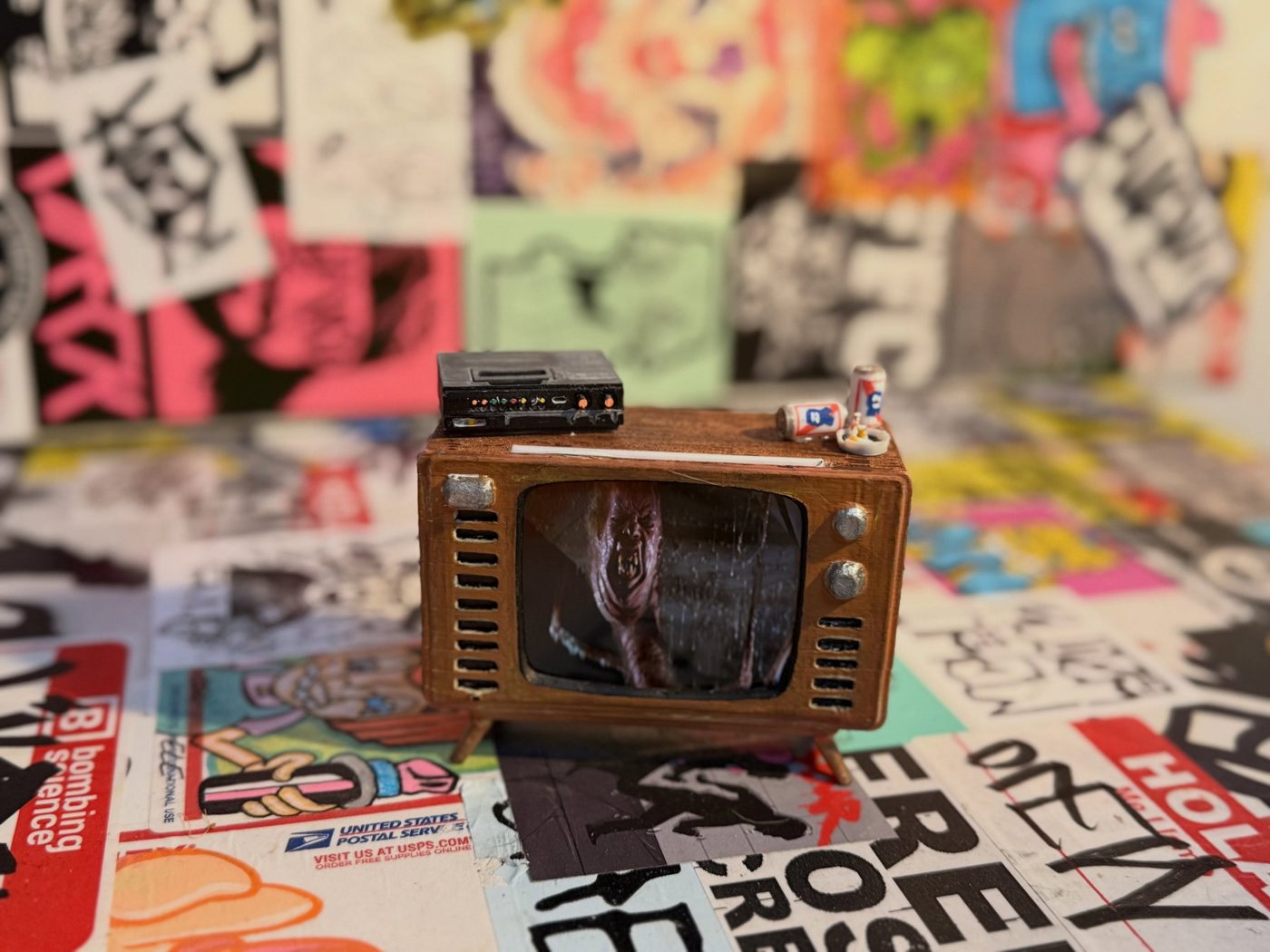 Tiny handmade wooden TV with a monster on screen, sitting on a sticker-covered surface